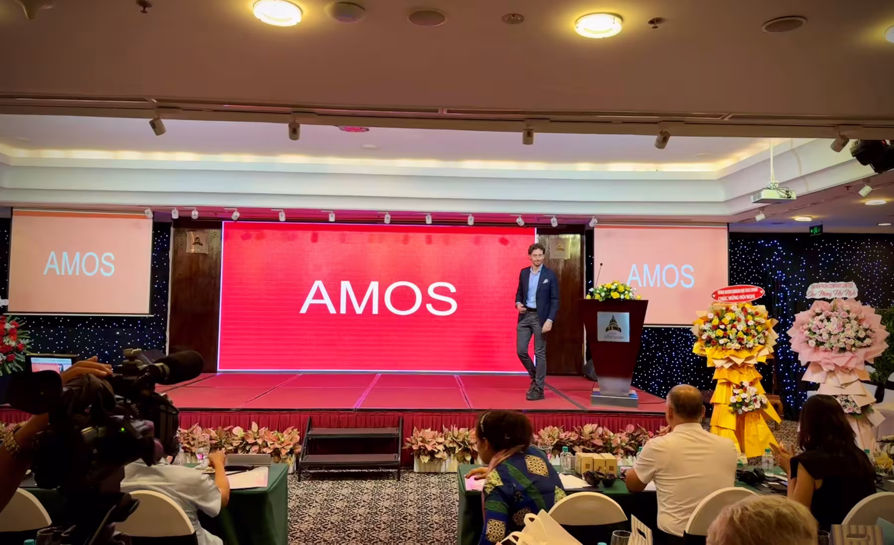 A man stands on a stage with a red screen behind him displaying the word "AMOS." People are seated at tables in front of the stage, with flower arrangements on either side.