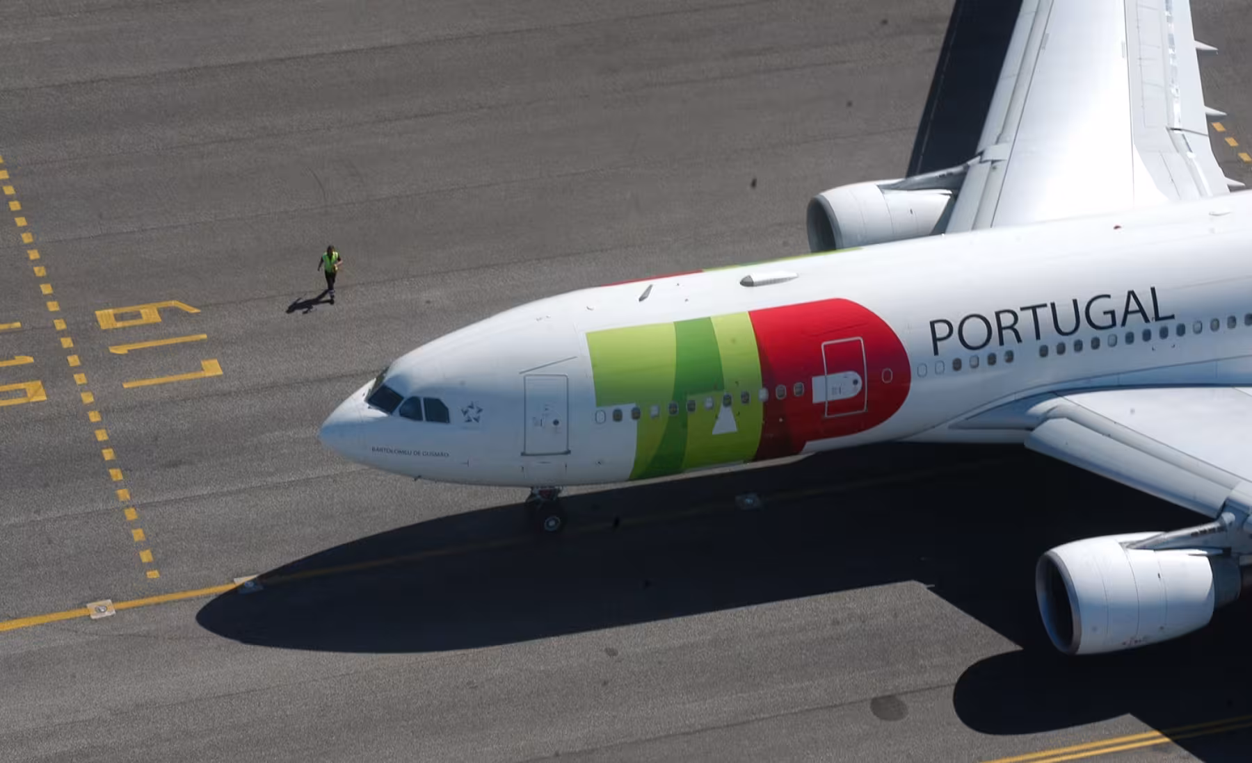 Top-down view of a TAP Air Portugal plane with a red and green logo, taxiing on the runway. A worker in a reflective vest is visible on the left, walking along the concrete near the aircraft.