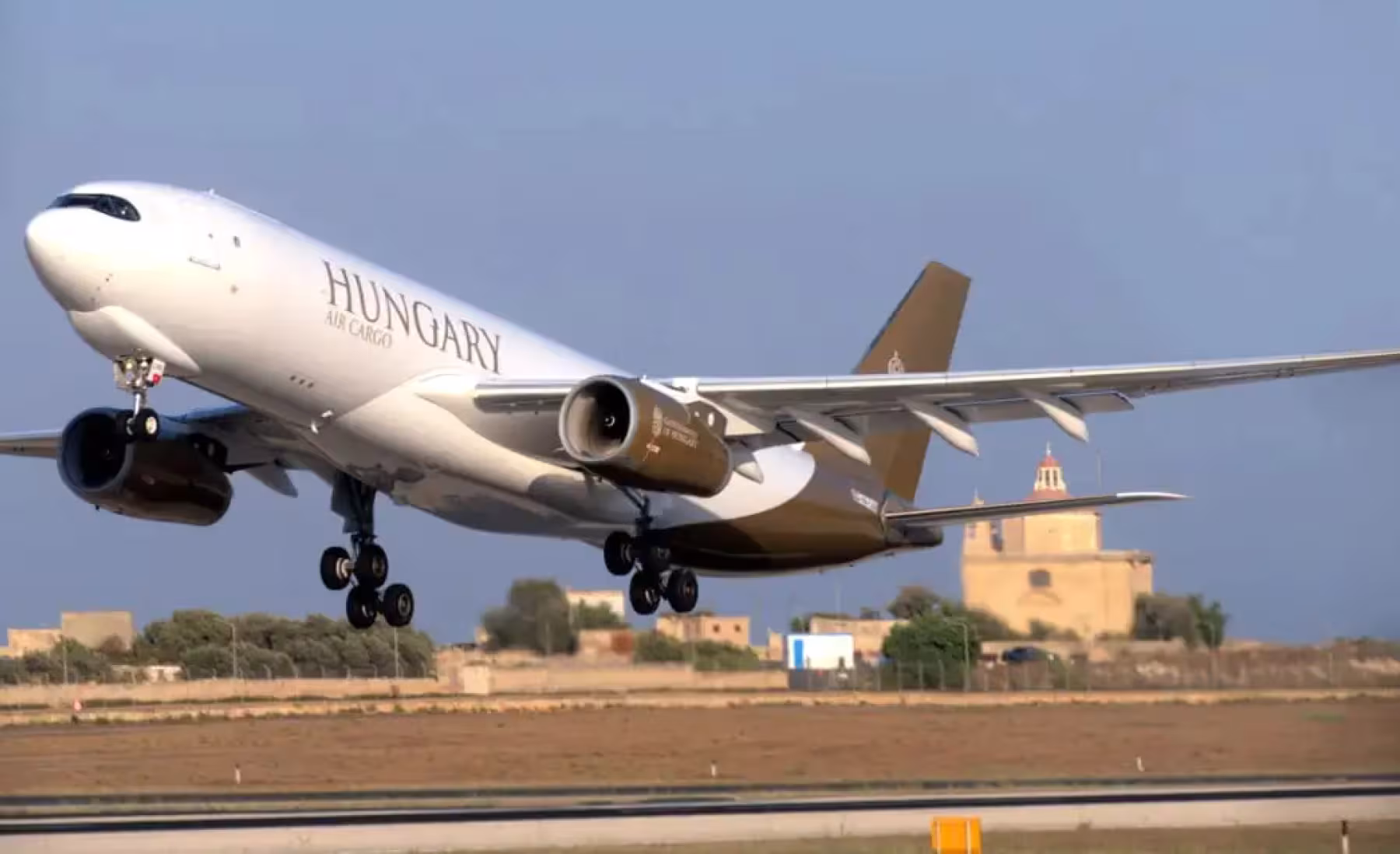 A Hungary-registered Airbus A330 takes off, with landing gear extended. The white fuselage features "HUNGARY" and gold-brown accents. The Hungarian flag colors appear on the winglets, with a historical building in the background.