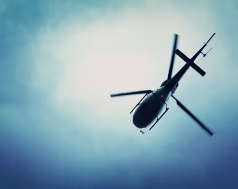 Helicopter seen from below in flight. Dark silhouette with spinning rotor blades against a cloudy blue sky.