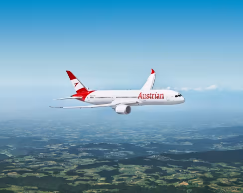Austrian Airlines passenger aircraft in flight against a clear blue sky, viewed side-on, with red tail and engine accents above a green patchwork landscape below.