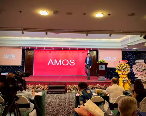 A man stands on a stage with a red screen behind him displaying the word "AMOS." People are seated at tables in front of the stage, with flower arrangements on either side.