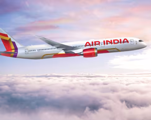 Air India airplane in flight, cruising over a sea of white clouds with a serene sky tinged in pink and blue hues. The aircraft&rsquo;s red-and-orange tail and engine accents stand out against the pale sky.