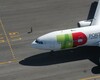 Top-down view of a TAP Air Portugal plane with a red and green logo, taxiing on the runway. A worker in a reflective vest is visible on the left, walking along the concrete near the aircraft.